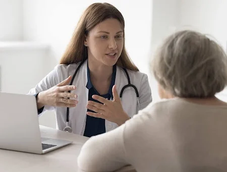 Positive doctor talking to elderly patient at office workplace, speaking, smiling with hands gestures, meeting with old lady at table with laptop, giving medical help.