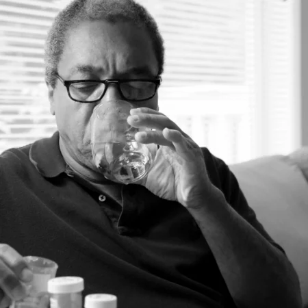 Black and white photo of Black man taking medication for bronchiectasis with a glass of water.